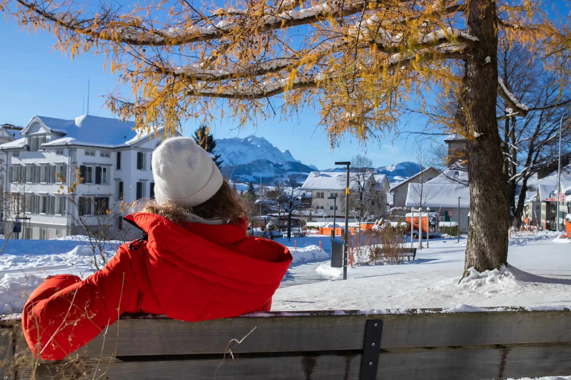 Person mit weißer Mütze und orange-roter Winterjacke sitzt auf einer Parkbank in einer Schneelandschaft und schaut auf das Hauptgebäude der AlpsteinClinic